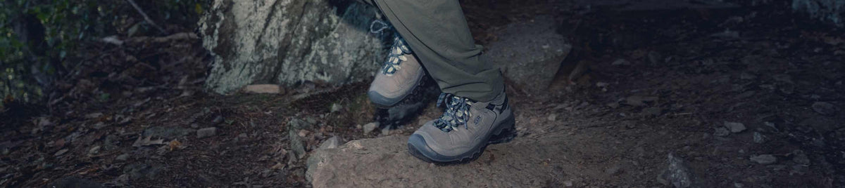 Eine lachende Frau in rosa T-Shirt, brauner kurzer Hose, Outdoorhut und braunen Wanderstiefeln hockt auf einem Felsen in bergiger, grüner Landschaft.