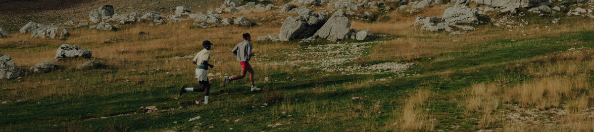 Zwei Trailrunner laufen über eine steinige Wiesenlandschaft im Gebirge, Fokus auf Bewegung, Tempo und Outdoor-Running.
