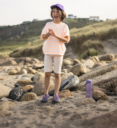 Kleines Kind mit lila Hut, lila Wasserflasche und lila Newport Boundless-Sandalen steht am Strandufer mit Felsen im Hintergrund