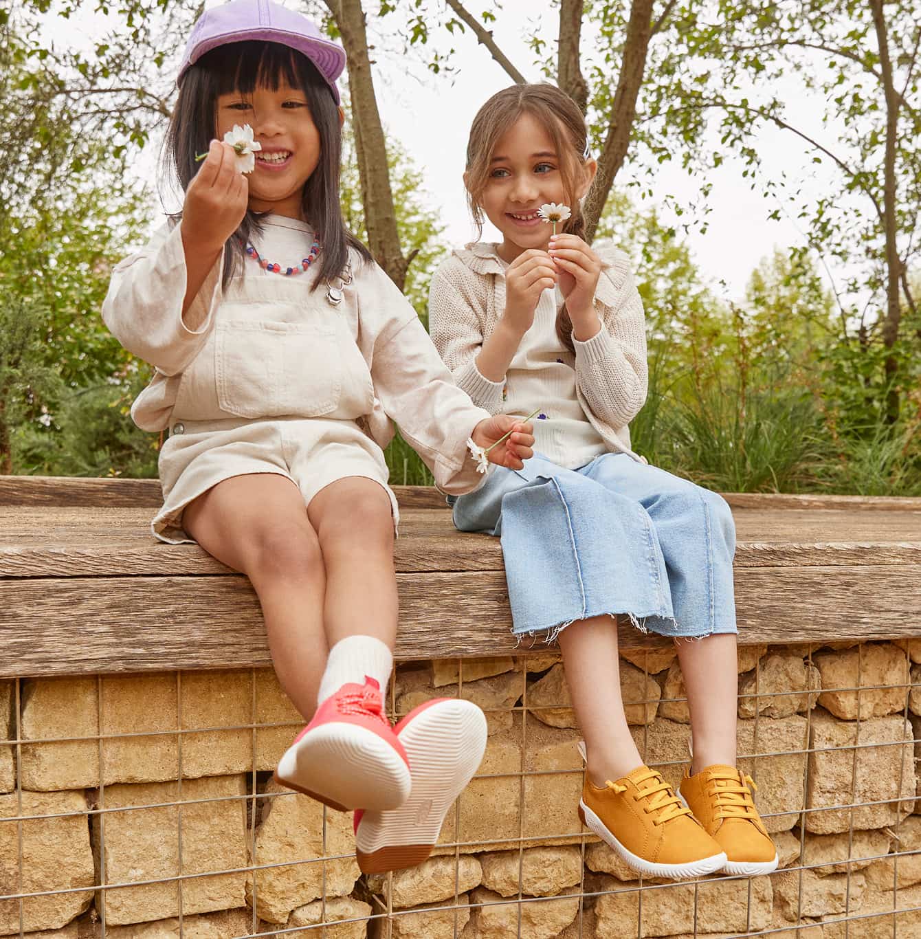 Zwei Mädchen in Kinderschuhen sitzen auf Holzbank mit Gänseblümchen - bunte KEEN KNX Sneaker und gelbe Schnürschuhe für Kinder im Frühling