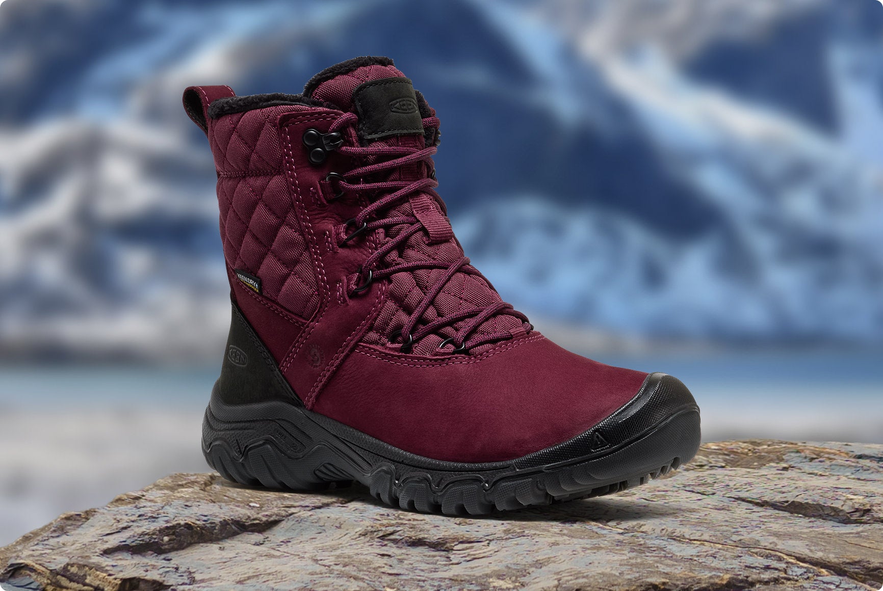 Weinroter KEEN Damen-Winterstiefel steht auf Fels vor verschneiter Bergkulisse.