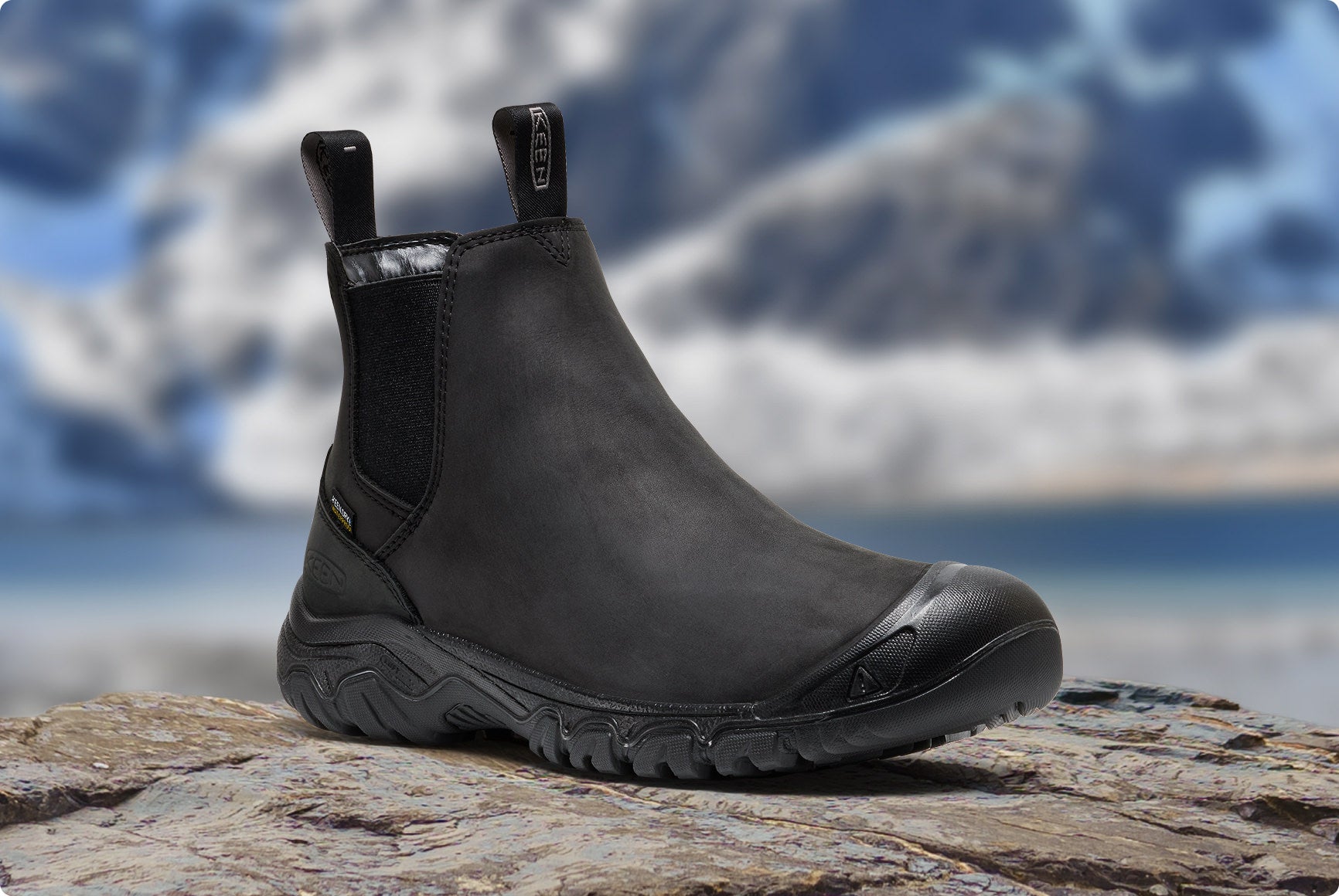 Schwarzer KEEN Herren-Winterstiefel steht auf Fels vor verschneiter Bergkulisse.