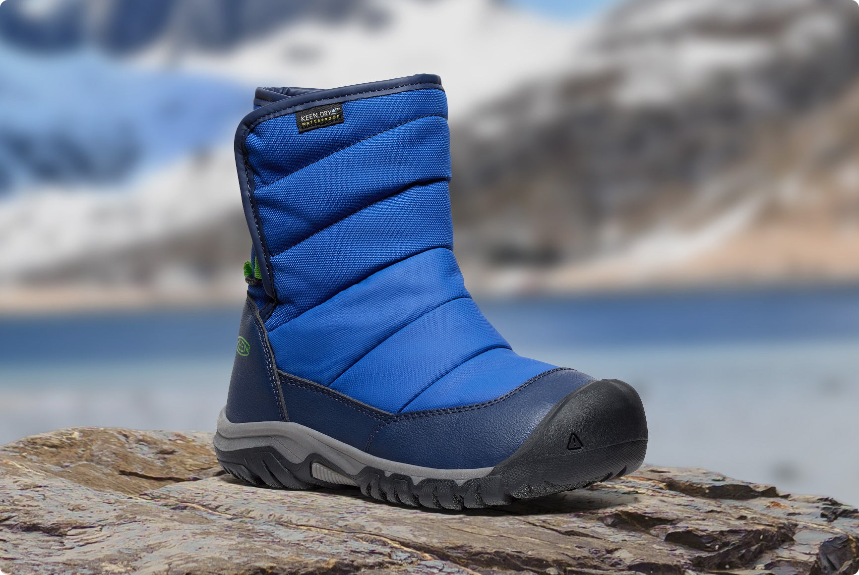 Blauer KEEN Kinder-Winterstiefel steht auf Fels vor verschneiter Bergkulisse.