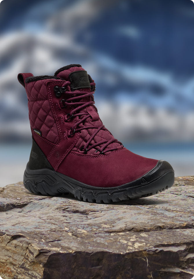 Weinroter KEEN Damen-Winterstiefel steht auf Fels vor verschneiter Bergkulisse.