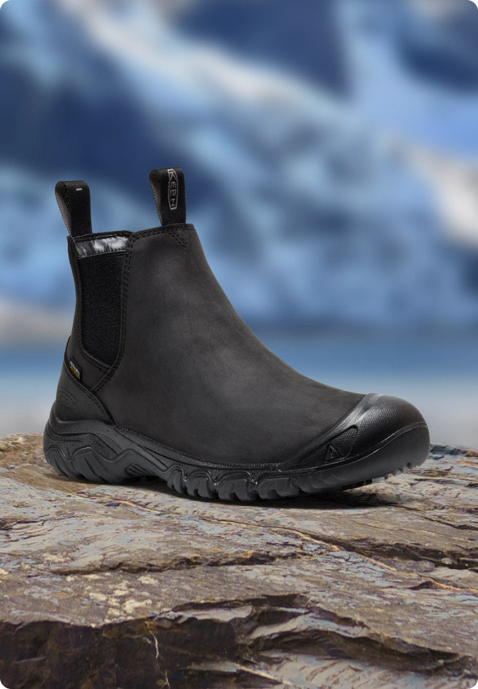 Schwarzer KEEN Herren-Winterstiefel steht auf Fels vor verschneiter Bergkulisse.
