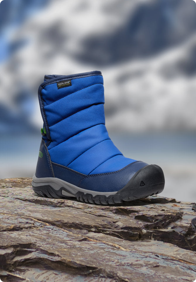 Blauer KEEN Kinder-Winterstiefel steht auf Fels vor verschneiter Bergkulisse.