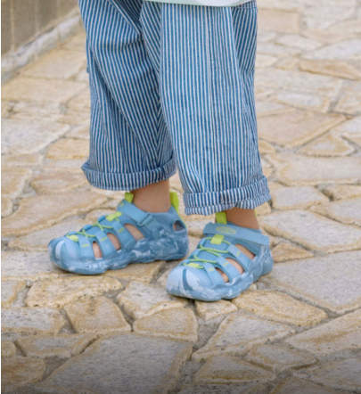 Niederknieender Blick auf einen kleinen Jungen in blauen Hosen, der blaue Hyperport-Wassersandalen trägt und auf den Fliesen sitzt