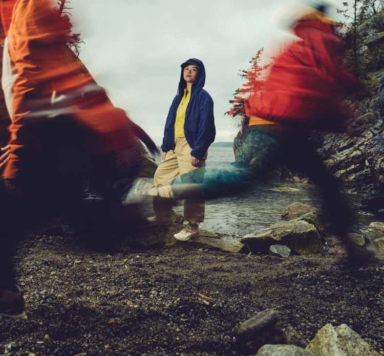 Person in Outdoor-Kleidung steht auf Felsen am Wasser während andere Personen in Bewegungsunschärfe vorbeigehen