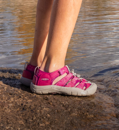 Knee-down shot of little girl wearing bright pink and gray Newport H2 sandals  while standing near waters' edge.