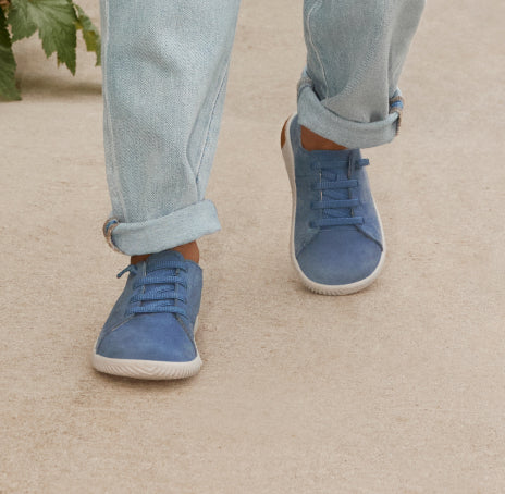 Niederknieender Blick auf einen kleinen Jungen in Denim, der blaue Leder-KNX-Sneaker trägt und auf hellbraunem Gehweg geht
